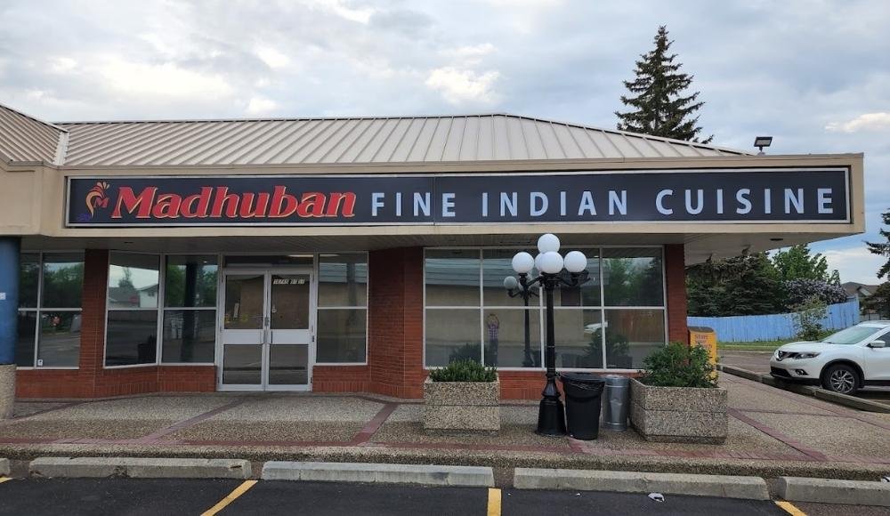 Storefront of Madhuban Fine Indian Cuisine features a large sign, expansive glass windows, and a spacious parking lot. Known for offering delightful buffets in Edmonton, this restaurant invites you to experience authentic Indian flavors in a warm and welcoming setting.