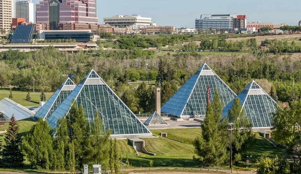 Four pyramid-shaped glass structures are situated in a green park area with a cityscape in the background. Trees and grass surround the pyramids, blending the natural and urban environments, making them one of the must-see attractions in Edmonton.