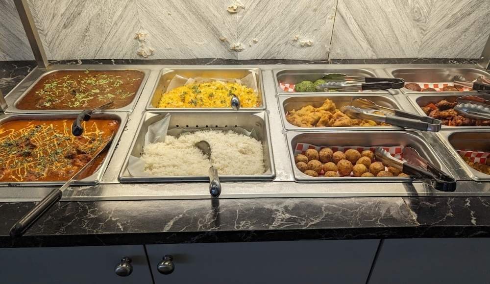 Buffet display with various hot dishes, including curry, rice, fried items, and vegetables in stainless steel trays on a marble counter—a true taste of buffets in Edmonton.