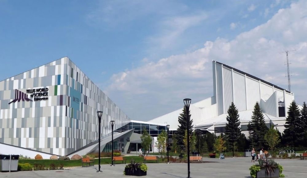 The image shows the Telus World of Science building in Edmonton, one of the top attractions in Edmonton Alberta, characterized by its modern architecture with geometrically patterned walls and a pedestrian area with benches and trees in the foreground.