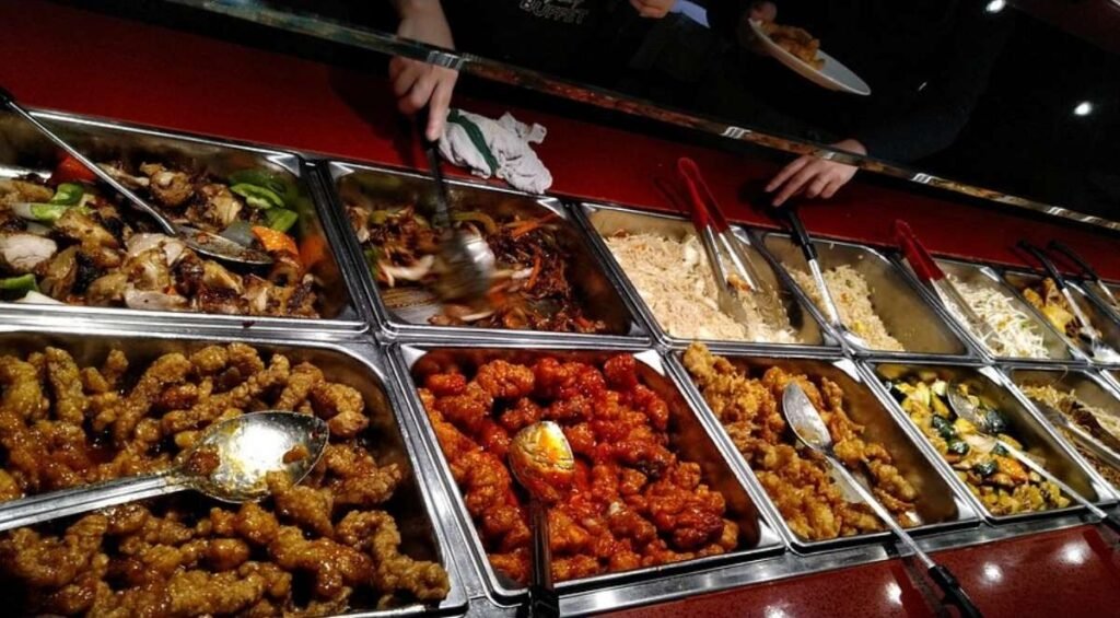 A buffet in Edmonton offers an array of dishes including fried foods, noodles, and vegetables, with serving spoons and tongs in each tray. Two people are serving themselves food.