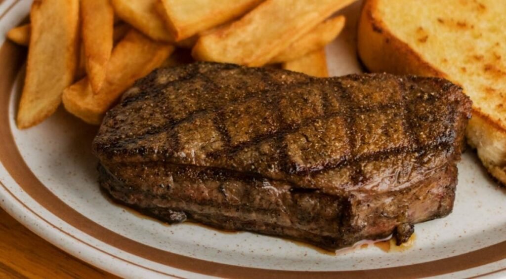 A grilled steak with crisscross marks, reminiscent of the finest steakhouses in Edmonton, is accompanied by thick-cut fries and a slice of toasted bread on a tan and white plate.