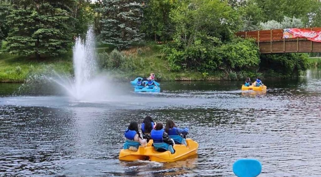 At Bower Ponds, people in colorful pedal boats navigate a serene pond with a sparkling fountain. Majestic trees and a charming wooden bridge provide the perfect backdrop for this picturesque setting.