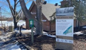 A sign at scenic Bower Ponds in Waskasoo Park, Red Deer, stands next to a charming brick building amidst bare trees and a snow-blanketed landscape.