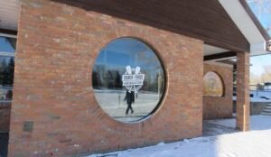 Large circular window with the "Bowin Ponds Recreation" logo graces a brick building. Snow and trees, reminiscent of the serene landscapes at Bower Ponds, are beautifully reflected in the glass.