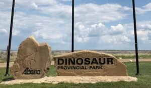 A carved dinosaur stands proudly beneath a cloud-filled sky on the sign for Dinosaur Provincial Park, Alberta.