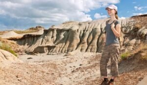 A person wearing a cap and camouflage pants stands smiling in the rocky, desert-like landscape of Dinosaur Provincial Park under a partly cloudy sky.