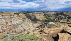 The rugged, rocky canyon landscape of Dinosaur Provincial Park features stratified cliffs, sparse vegetation, and a cloudy sky in the background.