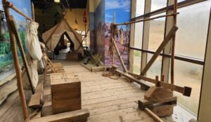 A wooden walkway with mining equipment and a canvas tent, surrounded by large murals of landscapes, inside a museum exhibit reminiscent of Dinosaur Provincial Park.