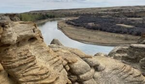 A river winds through the rocky expanse of Writing-on-Stone Provincial Park, showcasing intricate layered formations in the foreground. Sparse vegetation hugs the banks under a cloudy sky, adding to the mystique of this natural wonder.