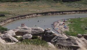 Visitors unwind and swim along a riverbank at Writing-on-Stone Provincial Park, where unique rock formations grace the foreground and lush grassy fields stretch into the background.