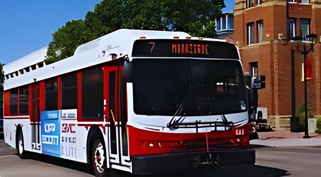 A city bus, part of the Red Deer Transit system, with route number 7 to Hillgate, is on the street near a red brick building and trees.