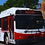 A city bus, part of the Red Deer Transit system, with route number 7 to Hillgate, is on the street near a red brick building and trees.