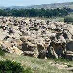 A stunning landscape at Writing-on-Stone Provincial Park showcases numerous sandstone rock formations, lush green vegetation, and a serene river under a clear blue sky. It's essential to know the area's natural beauty is also steeped in rich cultural history.