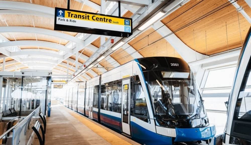 A modern light rail train, part of the Edmonton Transit Service, is stopped at a station platform under a wooden arched roof. A sign above reads "Transit Centre South Park & Ride.