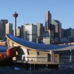 Visit Calgary to witness its stunning skyline, where modern skyscrapers tower alongside the distinctive Saddledome arena, all set against a clear blue sky.