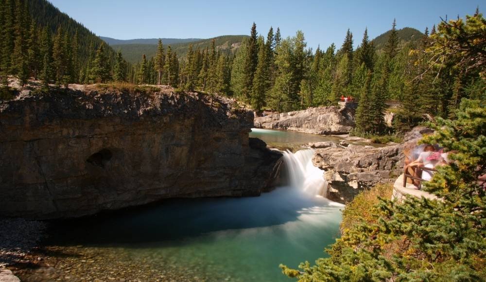 A small waterfall flows into a turquoise pool surrounded by rocky cliffs and dense evergreen forest under a clear blue sky. A person stands on a nearby viewpoint, capturing the serene beauty often found while wilderness camping in Alberta's breathtaking landscapes.