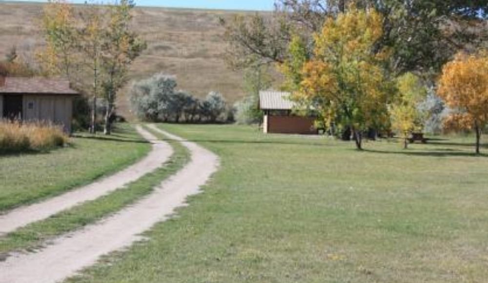 A dirt path meanders through a grassy area with trees and a small building in the background on a clear day, evoking the charm of camping near Calgary.