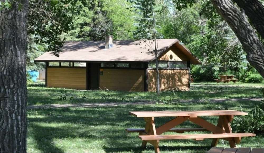 Nestled among towering trees, this quaint wooden cabin with a sloped roof invites you to experience adventure camping in Alberta. A nearby wooden picnic table on the grassy expanse completes this idyllic hideaway, perfect for those seeking tranquility and natural beauty.