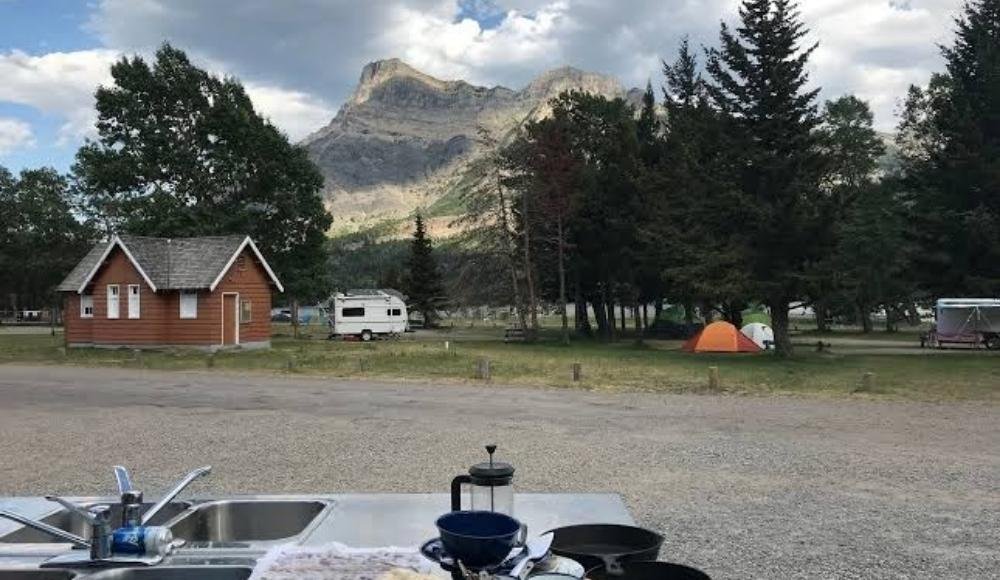 Nestled amidst the trees and mountains, this Alberta campsite offers a rustic cabin, RV, and tents. The picnic table is set with a camping stove, dishes, and a French press—perfect for an adventure camping experience in the heart of wilderness camping Alberta.