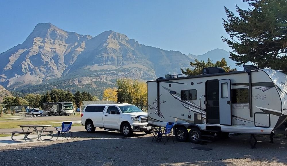 White SUV and camper trailer parked in a scenic mountainous campground at Waterton National Park, with a picnic table and chairs set up nearby under a clear sky.