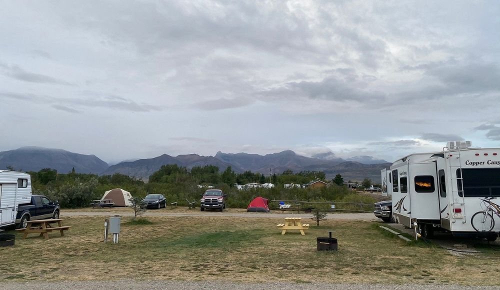 RV park scene with campers, trailers, picnic tables, and parked vehicles under a cloudy sky. Mountains are visible in the background, offering a serene setting perfect for outdoor activities. Consider these camping tips for an unforgettable experience at Waterton National Park.