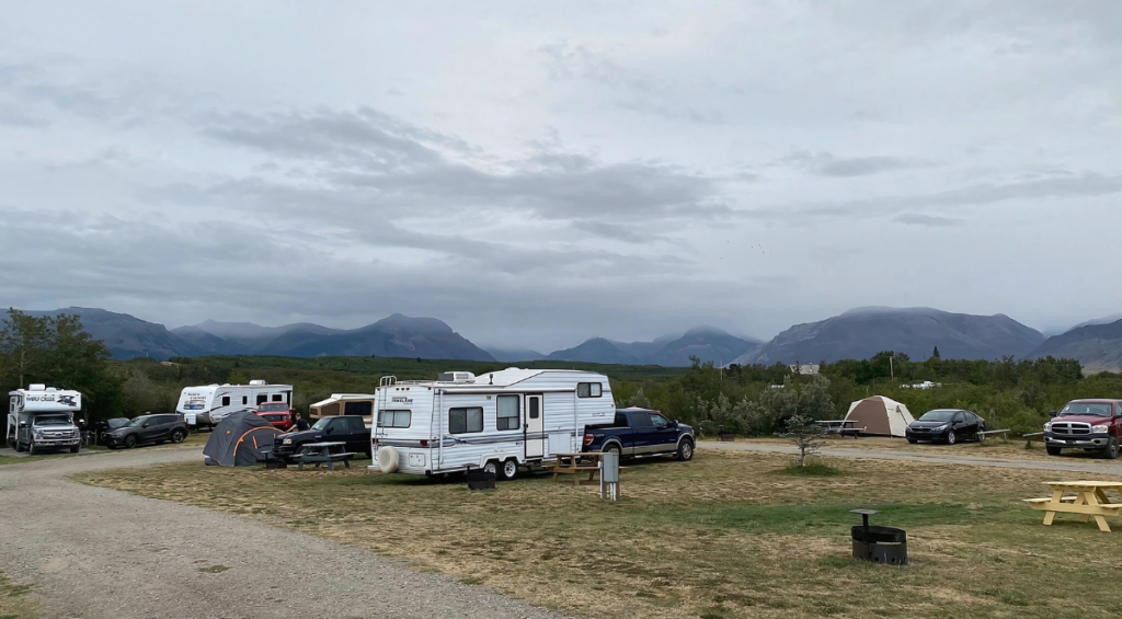 A campground scene with several RVs, cars, and tents dotting the grassy terrain, perfect for nature adventures. Mountains stand majestically in the background under cloudy skies—ideal for those seeking camping tips and outdoor activities.