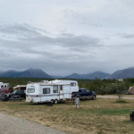 A campground scene with several RVs, cars, and tents dotting the grassy terrain, perfect for nature adventures. Mountains stand majestically in the background under cloudy skies—ideal for those seeking camping tips and outdoor activities.