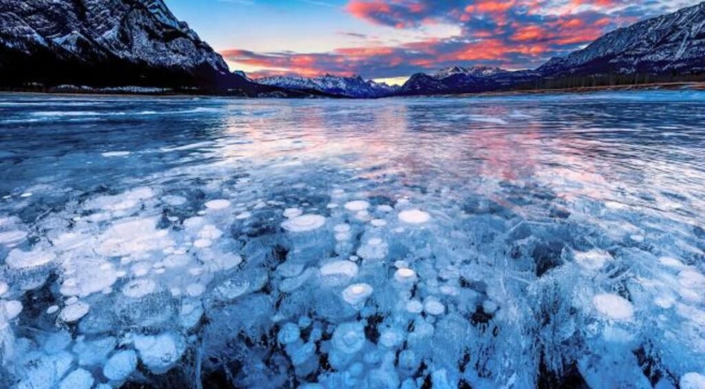 Frozen lake with visible ice bubbles beneath the surface, surrounded by snow-capped mountains at sunset with colorful clouds in the sky—an iconic scene at Abraham Lake.