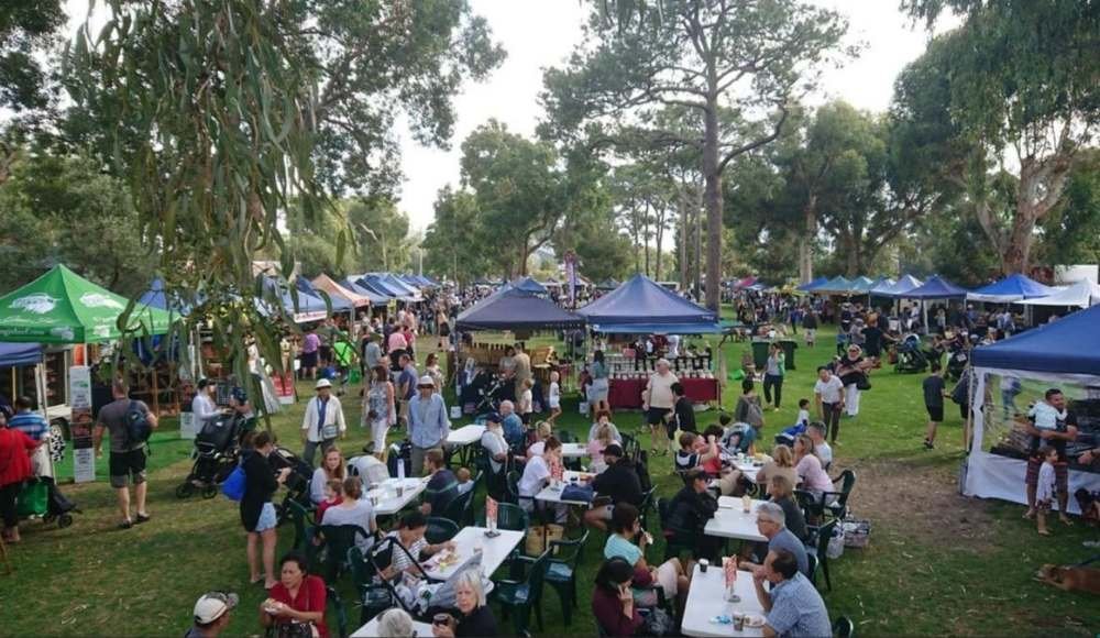 People strolling and relaxing among the colorful tents at an outdoor festival in a park with trees, as if enjoying a day at the Red Deer Farmers Market.