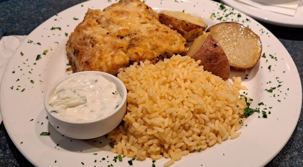 A plate with baked fish topped with a golden crust, served alongside potato wedges, yellow rice, and a small bowl of creamy sauce. Garnished with chopped herbs, this delightful dish is a testament to the culinary skills found in restaurants in Red Deer.