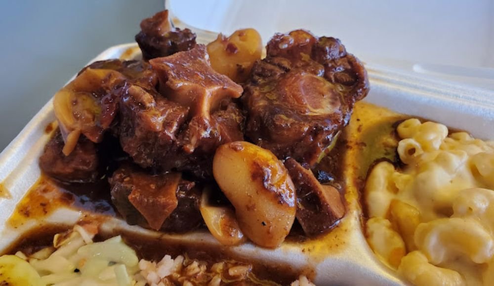 A serving of braised oxtail with potatoes in rich brown sauce nestles in a takeout container beside creamy macaroni and cheese, reminiscent of the comforting dishes you'll find at restaurants in Red Deer.