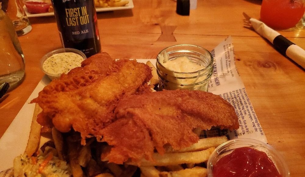 A plate of fish and chips, surrounded by an array of condiments and complemented by a red ale beer, graces this wooden table—a true taste sensation found in the best restaurants in Red Deer.