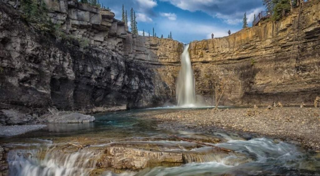 Waterfall cascades over a rocky cliff into a clear pool at Crescent Falls, Alberta, surrounded by steep stone walls and conifer trees under a partly cloudy sky.