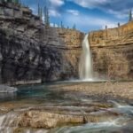 Waterfall cascades over a rocky cliff into a clear pool at Crescent Falls, Alberta, surrounded by steep stone walls and conifer trees under a partly cloudy sky.