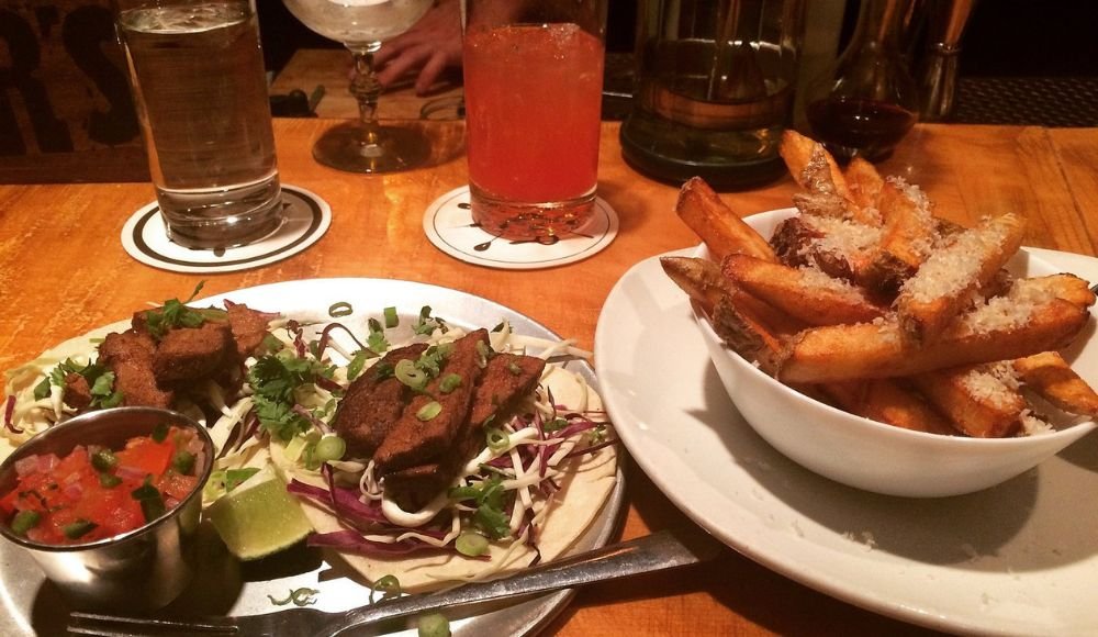 A plate with two tacos, salsa, lime wedge, and a bowl of seasoned fries topped with cheese sit invitingly at one of the popular restaurants in Red Deer. Accompanied by a refreshing glass of water and a vibrant red cocktail on coasters, it's the perfect dining experience on the wooden table.