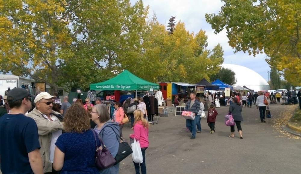 A bustling outdoor market with people strolling and browsing stalls under green trees at the Red Deer Farmers Market. Various tents and booths line the path, offering food and goods in a vibrant community setting.