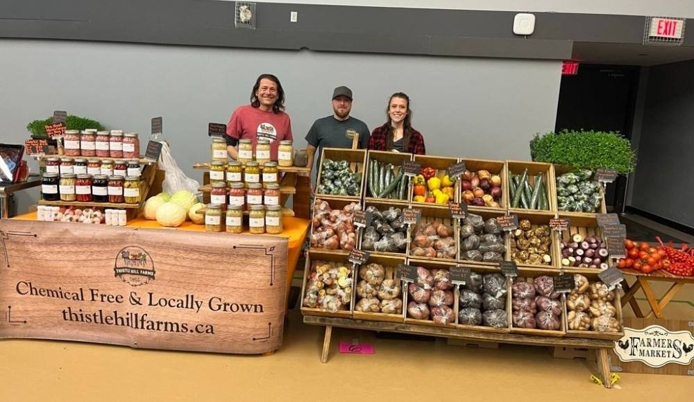 Three people stand behind a market stall at the Red Deer Farmers Market, showcasing an array of assorted vegetables and jars. The vibrant banner proudly declares "Chemical Free & Locally Grown," accompanied by a website URL.