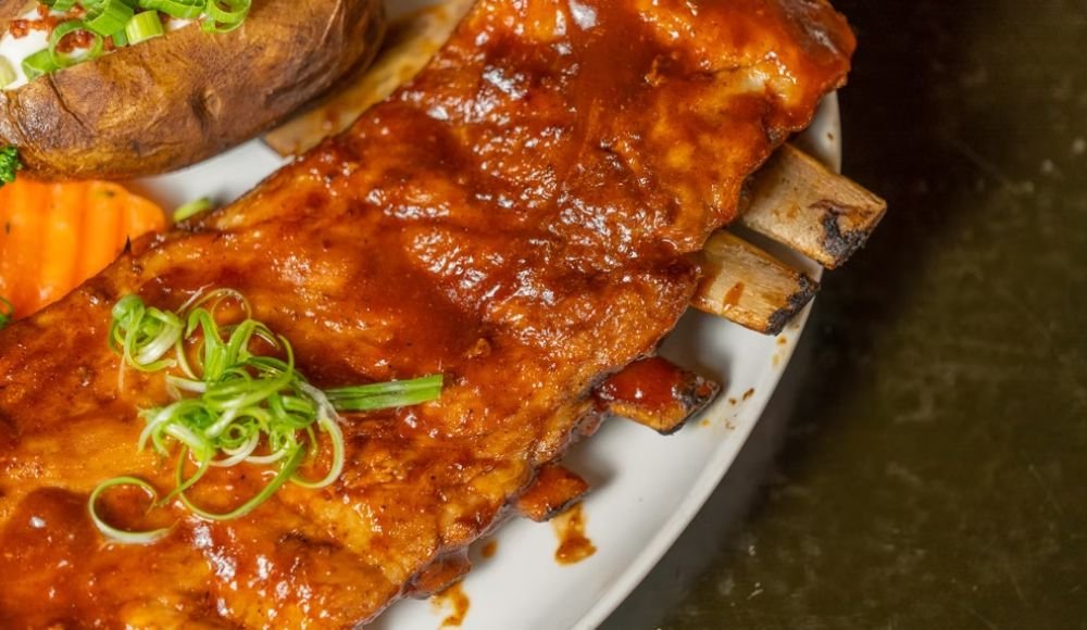 A sumptuous plate of cooked ribs glazed with barbecue sauce, garnished with green onions, and served alongside a baked potato and vibrant vegetables awaits you at one of the finest restaurants in Red Deer.