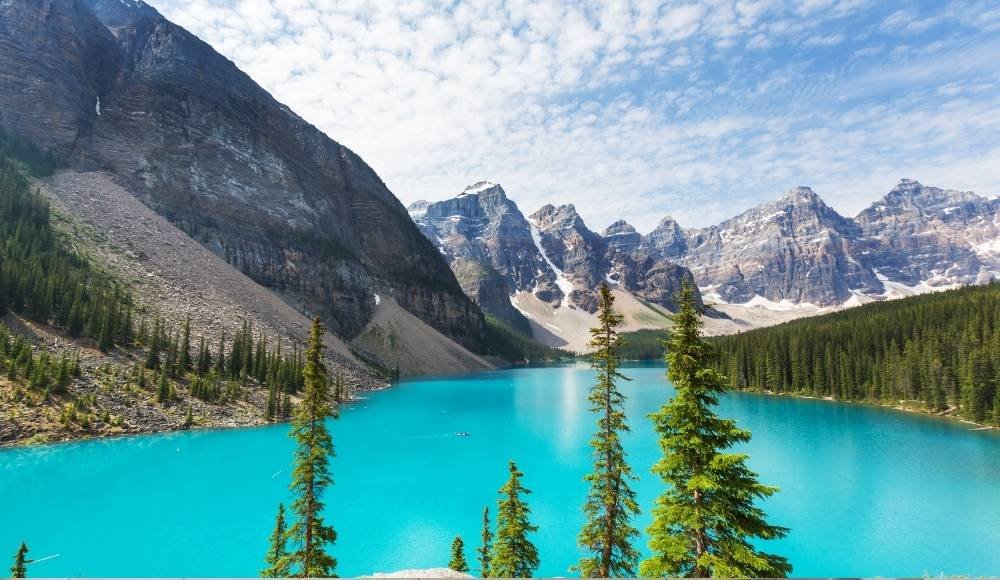 A turquoise lake, considered one of the best lakes in Alberta, is surrounded by pine trees, rocky slopes, and snow-capped mountains under a partly cloudy sky.