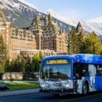A blue city bus drives past a large historic hotel with mountainous scenery and trees in the background, offering visitors a glimpse of How To Get To Banff National Park.