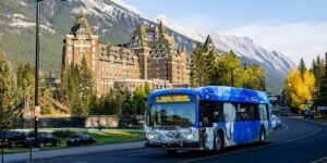 A blue city bus drives past a large historic hotel with mountainous scenery and trees in the background, offering visitors a glimpse of How To Get To Banff National Park.
