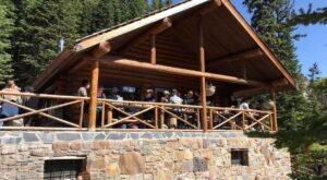 A group of people sit and stand on the wooden porch of a rustic log cabin, surrounded by trees and greenery, taking a break after completing the scenic Lake Agnes Tea House Trail.
