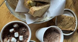 A basket with sandwich halves, two cups of hot chocolate (one with marshmallows), and a cookie on a dish, all placed on a wooden table—the perfect treat after completing the Lake Agnes Tea House hike.