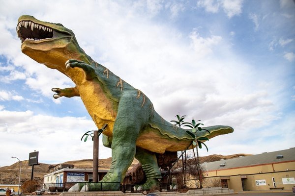 A large, green and yellow T. rex dinosaur statue stands outdoors beside artificial palm trees in Alberta, with buildings and hills in the background under a partly cloudy sky—one of the region’s top destinations for dinosaur enthusiasts.