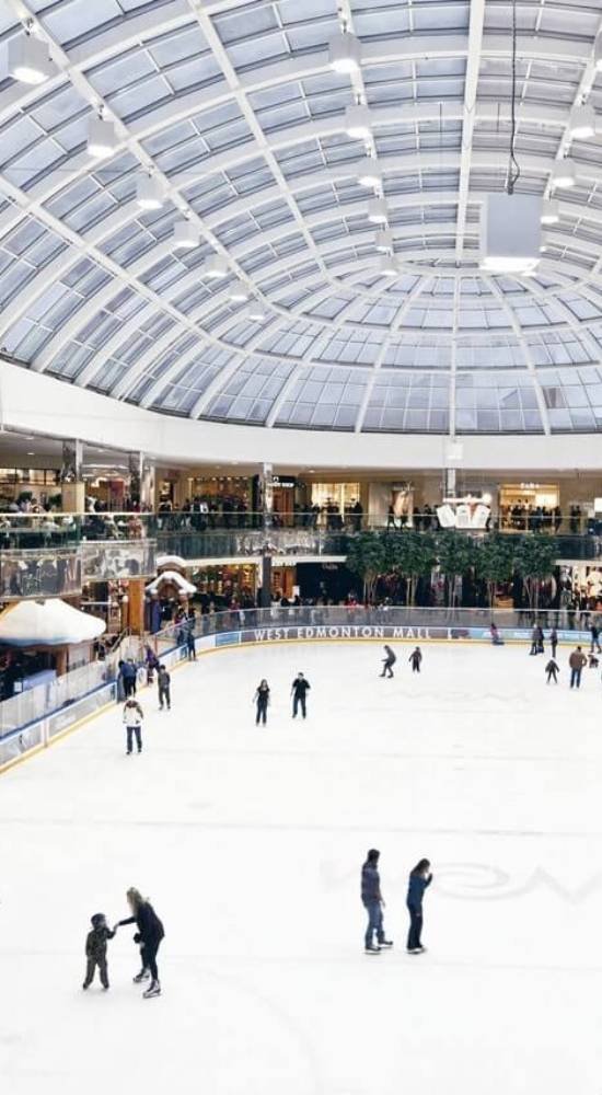 People skate on an indoor ice rink under a large glass dome at West Edmonton Mall; shops and spectators are visible in the background.