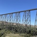 A tall steel train bridge extends across a valley with trees and buildings beneath it, set against a clear blue sky—a must-see highlight featured in many Lethbridge Alberta travel guides.