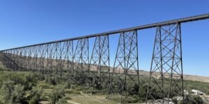 A tall steel train bridge extends across a valley with trees and buildings beneath it, set against a clear blue sky—a must-see highlight featured in many Lethbridge Alberta travel guides.