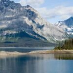 A calm lake with a small forested island, surrounded by tall, rugged mountains with snow patches under a partly cloudy sky—one of the breathtaking wonders in Alberta perfect for any Alberta travel guide.
