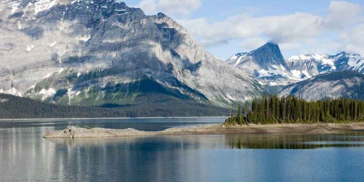 A calm lake with a small forested island, surrounded by tall, rugged mountains with snow patches under a partly cloudy sky—one of the breathtaking wonders in Alberta perfect for any Alberta travel guide.